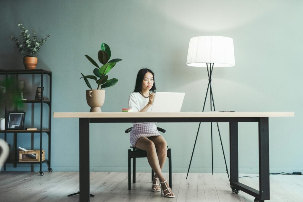 Asian woman working on a laptop in a modern office setting with a stylish lamp and plants.
